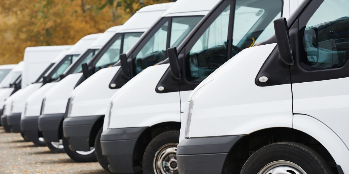 A row of white commercial auto vans parked in a lot, aligned side by side.